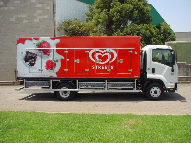 Delivery truck featuring vibrant red branding for Streets ice cream, showcasing images of strawberries and chocolate, parked on green grass.