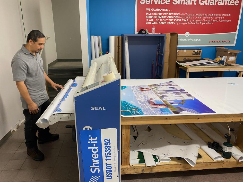 Man operating a wide-format printer in a design studio, preparing vibrant promotional materials for vehicles and businesses, featuring colorful graphics and printing equipment.